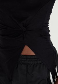 Schwarzes Oberteil aus weichem Stoff, mit einem verdrehten Knoten-Detail an der Seite und einem Schlitzsaum, kombiniert mit lockeren schwarzen Hosen.