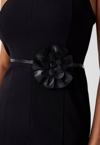 Schwarzes Kleid mit einem breiten schwarzen Ledergürtel, der mit einem großen, geschichteten Blumendetail versehen ist. Der Stoff hat eine glatte, matte Textur.