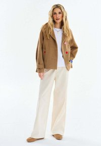 Žena s dlhými blond vlasmi, oblečená v hnedej bunde s farebnými gombíkmi, bielej košeli, širokých krémových nohaviciach a hnedých topánkach na bielom pozadí.