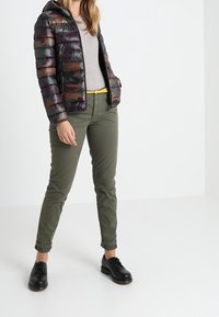 Puffjakke med multikolorede vandrette striber i mørkegrøn, brun og sort; parret med olivenfarvede bukser og sorte sko.