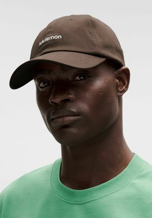 Detailní záběr osoby nosící hnědou čepici značky Lululemon a mátově zelené tričko s kulatým výstřihem, čelící dopředu s neutrálním výrazem.