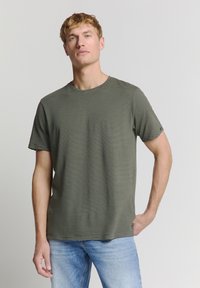 Olijfgroene T-shirt met korte mouwen, horizontale zwarte strepen, ronde hals en zachte textuur. Gemaakt in combinatie met lichtblauwe jeans, eenvoudig ontwerp.