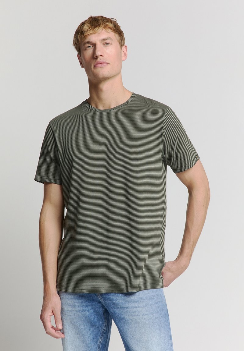 Olijfgroene T-shirt met korte mouwen, horizontale zwarte strepen, ronde hals en zachte textuur. Gemaakt in combinatie met lichtblauwe jeans, eenvoudig ontwerp.