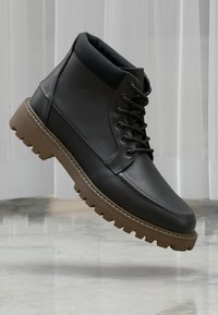 Botas de tobillo de piel negra con un cuello acolchado, suela de goma marrón y diseño con cordones. Presentan una superficie texturizada y costuras en contraste.