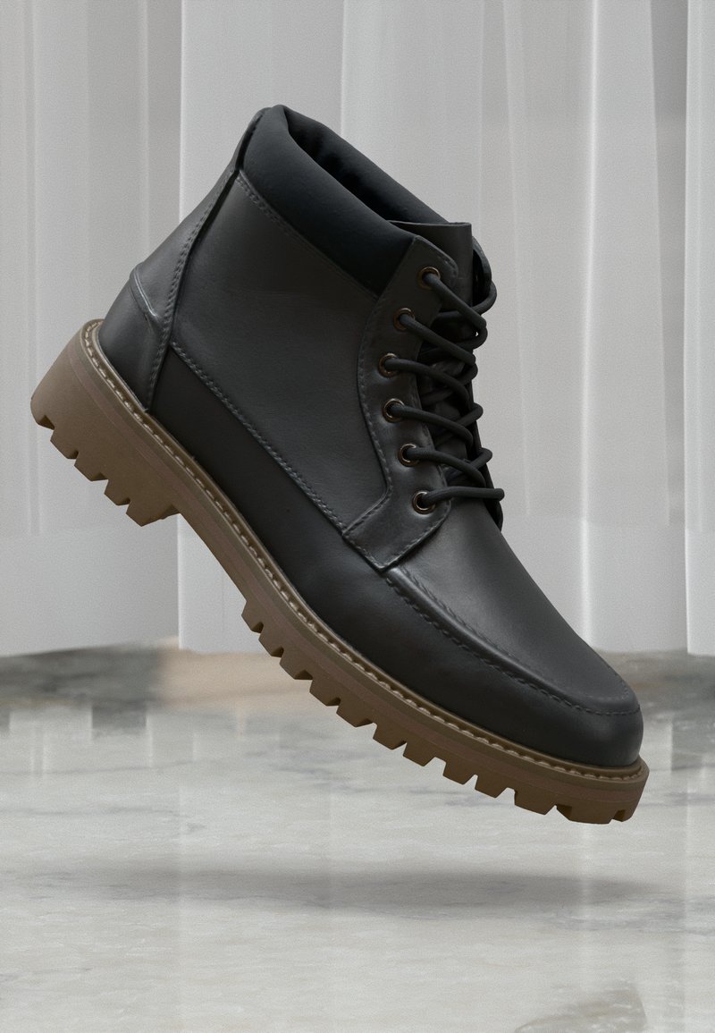 Botas de tobillo de piel negra con un cuello acolchado, suela de goma marrón y diseño con cordones. Presentan una superficie texturizada y costuras en contraste.