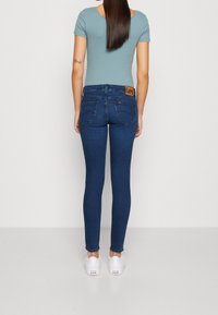 Jeans ajustados azules con una cintura de media altura y bolsillos traseros, con un parche de cuero marrón. Combinados con zapatillas blancas.