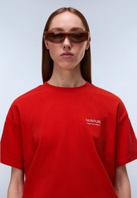 Κόκκινη βαμβακερή κοντομάνικη μπλούζα με λευκό κεντημένο λογότυπο στο αριστερό μέρος του στήθους. Το μοντέλο φορά καφέ οβάλ γυαλιά ηλίου.