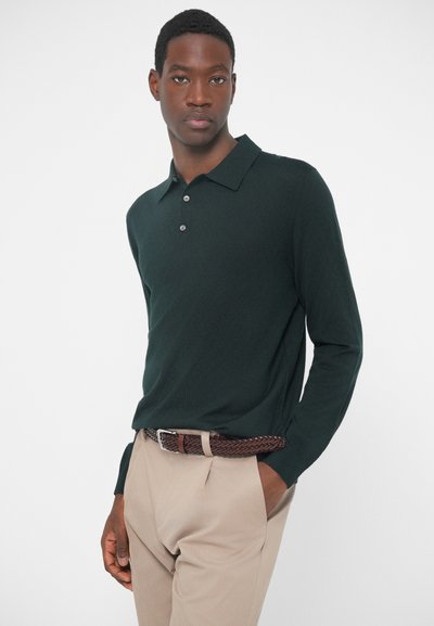 Σκούρο πράσινο πουκάμισο πόλο με μακριά μανίκια, γιακά και τρία κουμπιά, σε συνδυασμό με φωτεινά μπεζ παντελόνια και καφέ πλεκτή ζώνη.