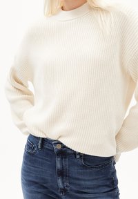 Pull en maille épaisse couleur crème avec un col montant et une coupe ample, associé à un jean en denim bleu foncé avec un bouton classique et une poche.