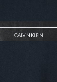 Marinblå bomullstyg med ribbad textur som har en svart etikett med "CALVIN KLEIN" tryckt i vita bokstäver över mitten.