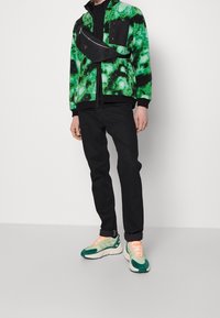 Giacca in pile verde e nera con motivo a zigzag, abbinata a jeans neri e sneakers verde e crema con dettagli arancioni. Presenta anche una borsa nera.