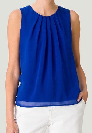 Bez rękawów królewska niebieska bluzka z plisowanym dekoltem, noszona z białymi spodniami, ręka swobodnie w kieszeni.