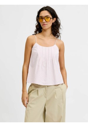 Femme aux cheveux bouclés mi-longs portant de grandes lunettes de soleil jaunes, un caraco blanc à fines rayures et un pantalon beige à jambes larges.