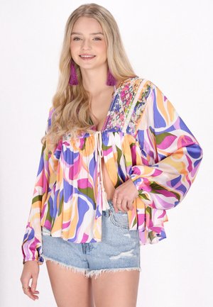 Jonge vrouw met lang blond haar draagt een kleurrijk gebloemde blouse met kwastje en spijkershort, glimlachend naar de camera.