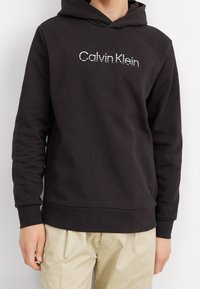 Sweat-shirt à capuche noir au toucher doux, arborant un logo blanc "Calvin Klein" sur le devant. Manches et ourlet côtelés.
