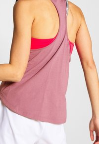 Světlé růžové tílko s kulatým lemem a otevřeným zadním dílem, vrstvené přes jasně růžovou sportovní podprsenku; vyrobeno z měkké, prodyšné tkaniny.