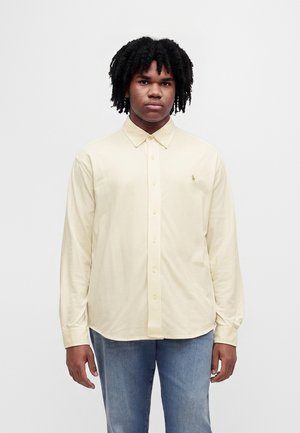 Jeune homme aux cheveux noirs bouclés portant une chemise jaune pâle boutonnée et un jean bleu, debout devant un fond blanc uni.