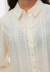 Femme portant un chemisier crème à boutons avec des rayures verticales en dentelle ajourée et un col pointu.