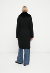Cappotto di lana nero con colletto in pelliccia texturizzata, vita contornata da una cintura e silhouette dritta. Indossato con jeans in denim a gamba larga e scarpe nere.