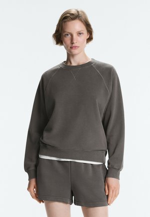 Kvinde iført en mørkegrå langærmet sweatshirt og matchende shorts, stående mod en ensartet lys baggrund.