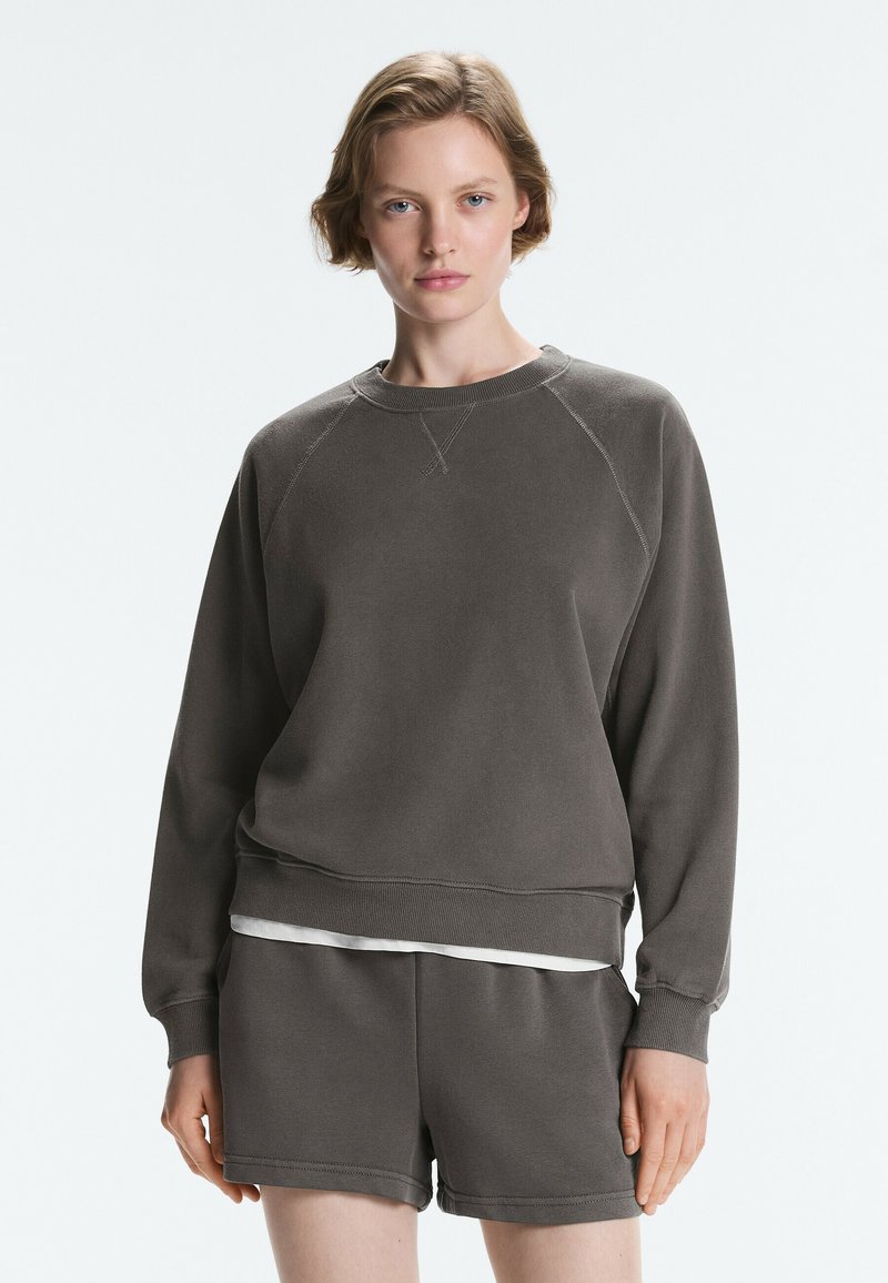 Femme portant un sweat-shirt gris foncé à manches longues et un short assorti, debout devant un fond clair uni.