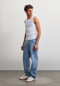 Homme debout en pose décontractée portant un débardeur blanc avec du texte, un jean bleu ample à cordon et des baskets blanches sur fond uni.