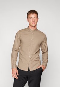 Camicia da uomo a maniche lunghe in tessuto beige con colletto alla cinese, dotata di chiusura frontale a bottoni e vestibilità aderente. Texture liscia, design minimalista.