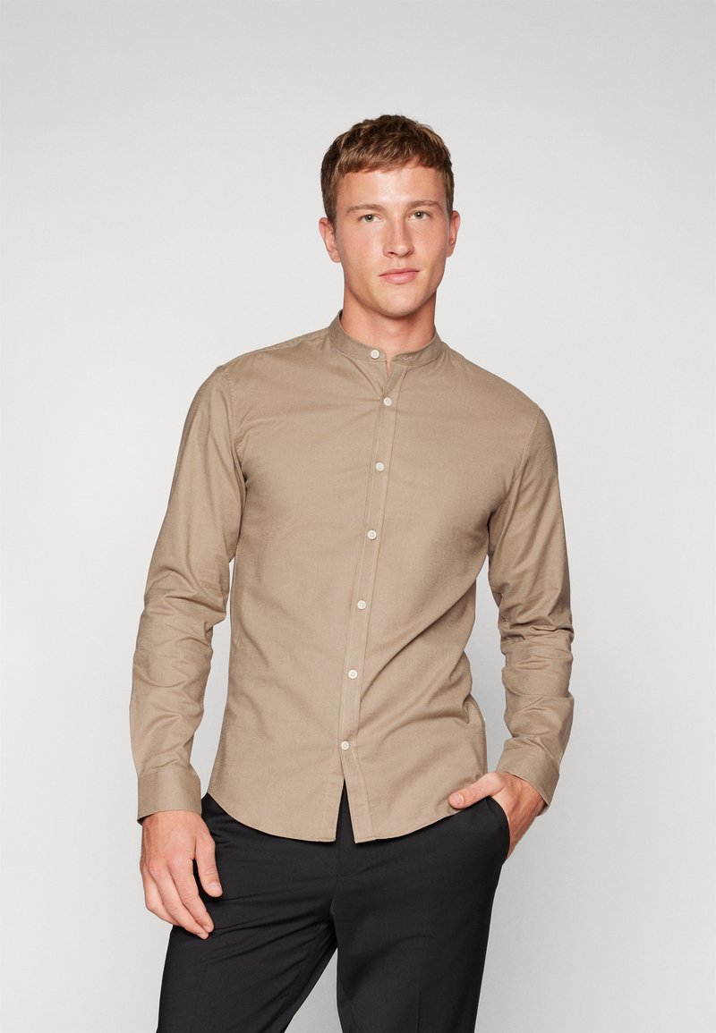 Camicia da uomo a maniche lunghe in tessuto beige con colletto alla cinese, dotata di chiusura frontale a bottoni e vestibilità aderente. Texture liscia, design minimalista.