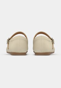 Μπεζ παπούτσια χωρίς κορδόνια με απαλό υφασμάτινο επάνω μέρος, λουράκι στον αστράγαλο και επίπεδη λάστιχη σόλα· διαθέτουν μικρή ασημένια αγκράφα στο λουράκι.