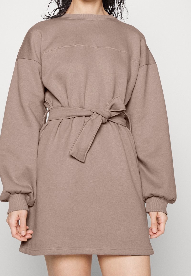 Robe-sweatshirt marron à manches longues avec une ceinture à nouer à la taille. Tissu doux, coupe décontractée, poignets côtelés et détails de couture sur les épaules.