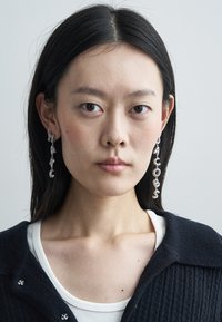 Ασημένια σκουλαρίκια με παιχνιδιάρικο σχέδιο γραμμάτων που σχηματίζουν τη λέξη "JACOBS", με σχέδιο αλυσίδας και λαμπερό φινίρισμα. Το μοντέλο φοράει πλεκτή μπλε ζακέτα.