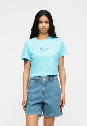 Jeune femme aux longs cheveux foncés portant un t-shirt court bleu clair avec "ROXY" et un short en jean, debout devant un fond uni.