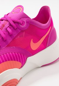 Světlé růžové sportovní boty s síťovaným svrškem, texturovanými akcenty a oranžovým logem swoosh. Hmotná bílá podrážka s jemnými designovými liniemi.