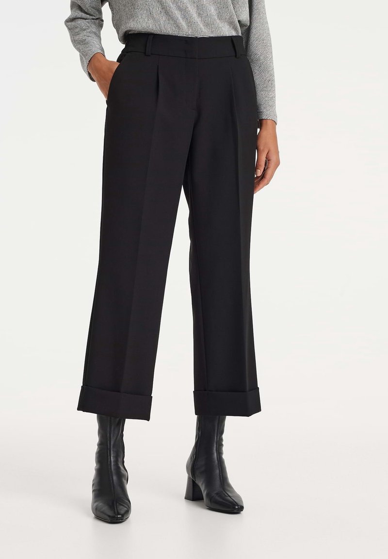 Personne portant un pantalon noir large et court avec des ourlets repliés, des bottines noires à talons et un haut gris clair à manches longues avec une main dans la poche.