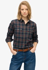 Navy and green plaid flannel shirt with long sleeves rolled up, featuring two chest pockets and a button-down collar. Worn with denim jeans and a brown belt.
