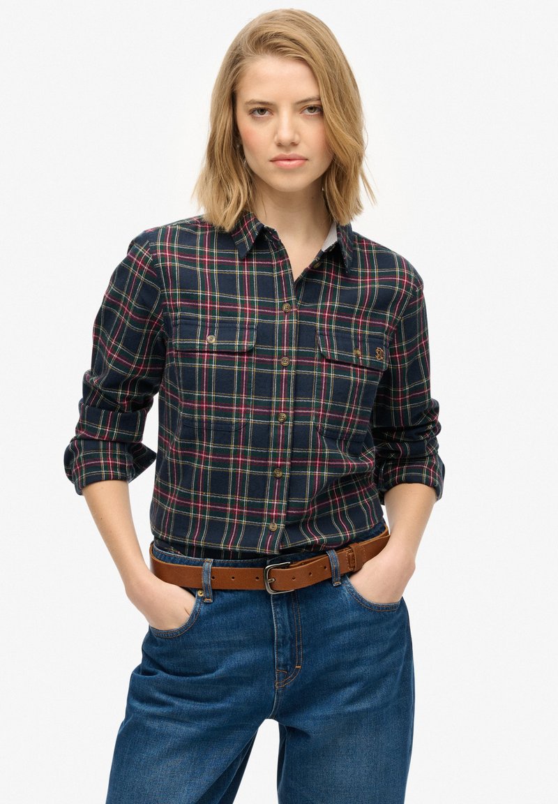 Navy and green plaid flannel shirt with long sleeves rolled up, featuring two chest pockets and a button-down collar. Worn with denim jeans and a brown belt.