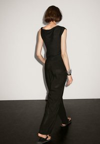 Black sleeveless top with draped neckline, paired with loose black pants and black sandals, viewed from the back against a neutral background.