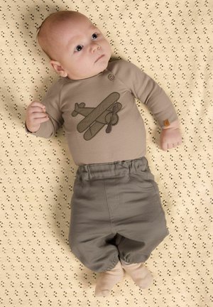 Baby som ligger på et mønstret beige teppe, iført en brun langermet skjorte med flydesign og grå bukser.