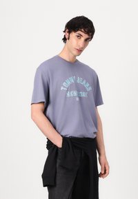 Trumpas rankovės, apvaliu kaklu purpurinis marškinėliai su mėlynu užrašu „TOMMY JEANS NEW YORK“, derinami su juodomis kelnėmis ir džemperiu, surištu aplink liemenį.