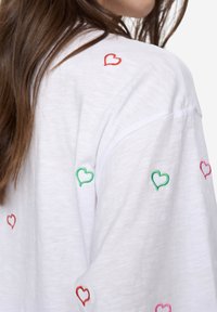 Bílá košile s krátkým rukávem a barevným výšivkou srdíček v červené, zelené a růžové barvě. Měkká textura a uvolněný střih.