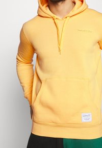 Żółta bluza z kapturem, wykonana z miękkiego materiału, z kieszenią kangurzą, ściągaczami przy rękawach oraz małym haftowanym logo. Widać widoczne detale maszyny szyjącej.