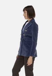 Blazer in velluto blu navy con design doppiopetto, bottoni dorati, rever a punta e tasche frontali, indossato con una maglietta bianca e pantaloni marroni.