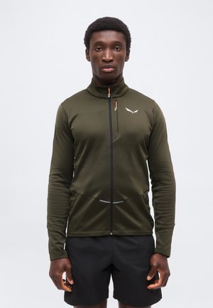 Mees seisab näoga ettepoole, kandes oliivirohelist lukuga jopet valge logoga ja musti lühikesi pükse ühevärvilise valge tausta ees.