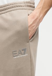 Beige jogger met elastische tailleband en zijzak, voorzien van geborduurd "EA7 Emporio Armani"-logo in contrasterende kleuren. Glad weefsel.