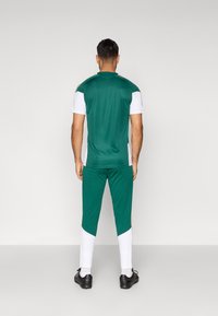 Vīrietis, ģērbies zaļā un baltā sporta apģērba komplektā ar melniem futbola apaviem, stāv vērsts prom pret vienkāršu balto fonu.