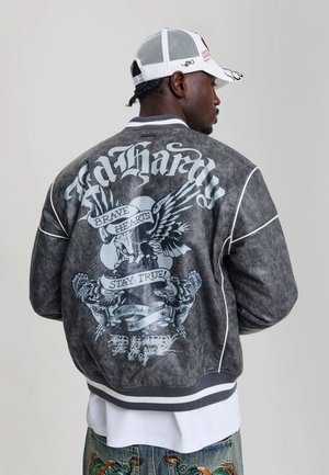 Mężczyzna noszący ciemną kurtkę Ed Hardy z wzorem orła i transparentu oraz białym tekstem, białą czapkę i haftowane dżinsy, widziany od tyłu.