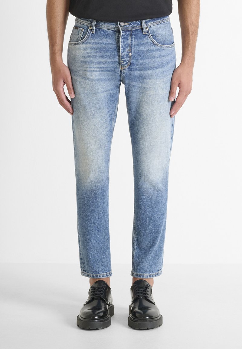 Jeans de mezclilla azul claro con un diseño deslavado, corte recto, estilo de cinco bolsillos y cierre de botón, combinados con zapatos de cuero negros.