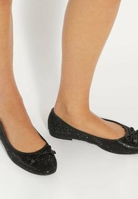 Zwarte balletschoenen met een ge textured, sprankelende bovenkant, afgeronde neus en strikdetail aan de voorkant. Platte zool voor gemakkelijke draagbaarheid.