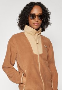 Brun fleecejacka med beige detaljer, hög krage och dragkedja. Har sidofickor och en diskret logotyp på bröstet.