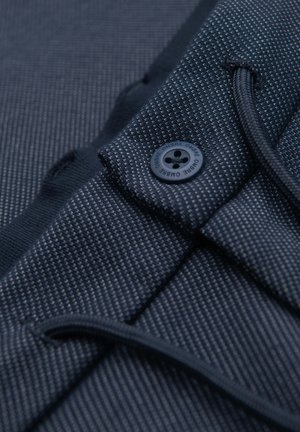 Detail tmavě modrých texturovaných kalhot s knoflíkem označeným "OMBRE" a viditelným stahovacím šňůrkou na elastickém pásu.